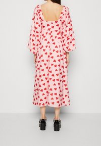 Kvinna klädd i en rosa midiklänning med röda hjärtan, puffärmar, fyrkantig öppen rygg och svarta platåskor, står vänd bort.