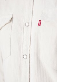 Gros plan sur le devant d'une chemise blanche en velours côtelé avec des boutons-pression métalliques et une étiquette rouge au logo Levi's sur la poche poitrine.