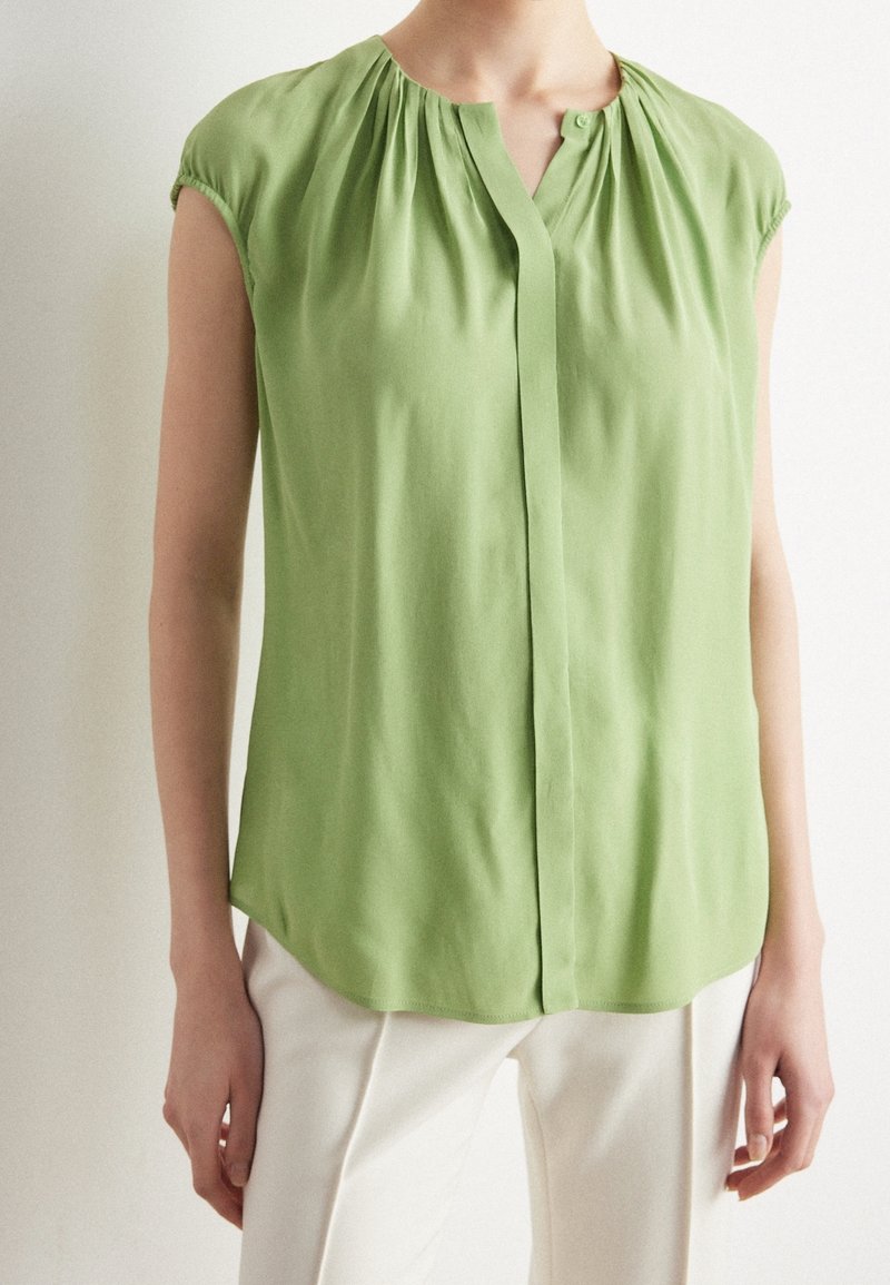 Femme portant un chemisier sans manches vert clair avec des plis au niveau du col et un pantalon blanc, posant devant un fond uni.