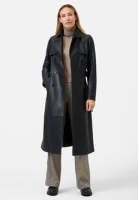 Black leather trench coat with double-breasted design, side pockets, and a collar. Worn over a beige turtleneck and patterned trousers.