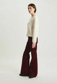 Pull crème à manches longues, ajusté, associé à un pantalon rouge foncé à jambes larges, texturé, avec un motif floral, porté avec des chaussures noires.