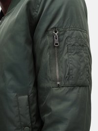 Gros plan d'une manche de veste verte avec une poche zippée verticale et des compartiments rectangulaires cousus sur le haut du bras.
