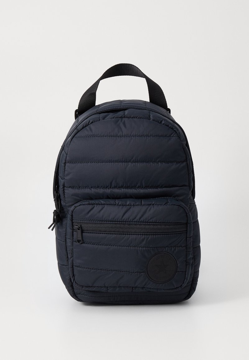 Petit sac à dos matelassé bleu marine avec une poche zippée à l'avant et une poignée supérieure noire sur fond blanc.