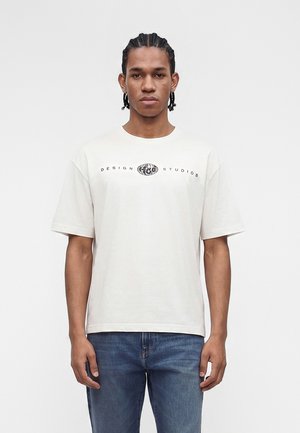 Άνδρας με κοτσίδες που φορά λευκό μπλουζάκι με το κείμενο «Design Studios» και λογότυπο γης, συνδυασμένο με μπλε τζιν, στέκεται μπροστά σε απλό φόντο.