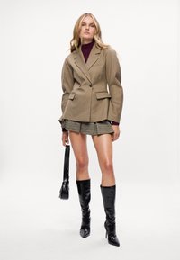 Blazer bege sob medida com ombreiras, bolsos com aba e um botão único. Combinado com uma saia mini xadrez e botas pretas brilhantes até ao joelho.