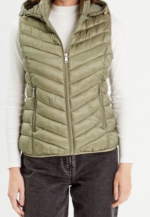 Veste matelassée sans manches vert olive avec fermeture éclair devant et poches zippées portée sur un top blanc côtelé à manches longues et un jean foncé.