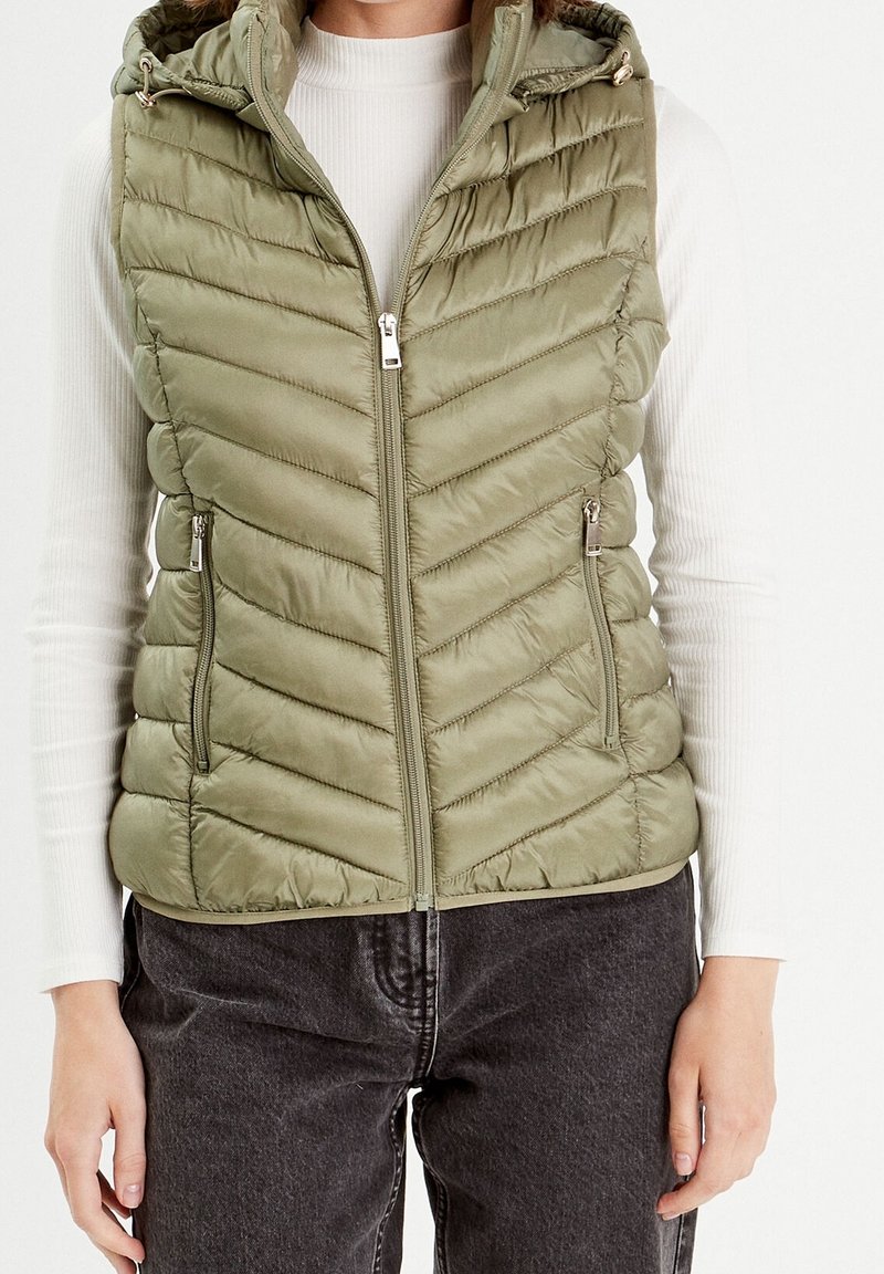 Veste matelassée sans manches vert olive avec fermeture éclair devant et poches zippées portée sur un top blanc côtelé à manches longues et un jean foncé.