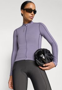 Lilla cykeltrøje med lange ærmer, lynlås foran og kontrasterende syninger, kombineret med sorte cykelshorts og en sort cykelhjelm.
