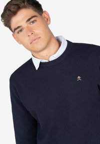 Granatowy sweter z dzianiny o teksturowanym wykończeniu, okrągłym dekolcie i małym, metalicznym logo na klatce piersiowej. Nałożony na jasnoniebieską koszulę z kołnierzykiem.