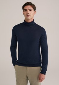 Hombre vestido con un suéter de cuello alto azul marino y pantalones beige, de pie con una mano en el bolsillo frente a un fondo liso.