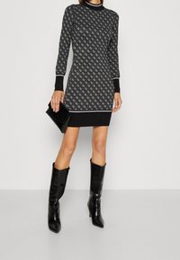 Robe en tricot à motifs noir et blanc, manches longues, col montant et poignets côtelés ; associée à des bottes noires à talons montant jusqu'aux genoux et une pochette.