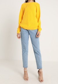 Żółty prążkowany sweter z szerokim dekoltem, połączony z jasnoniebieskimi jeansami zwężanymi oraz beżowymi sandałami na wysokim obcasie. Prosty design i faktura.