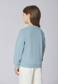 Bambino con lunghi capelli castani, vestito con una felpa azzurra chiara e pantaloni bianchi, in piedi rivolto di spalle su uno sfondo semplice.