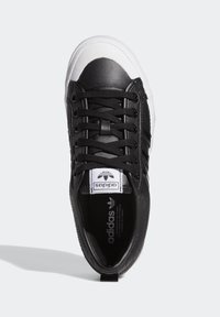 Černé kožené tenisky s bílým gumovým špičkovým krytem, černými tkaničkami a perforovanými bočními detaily. Mají etiketu na jazýčku s logem a texturovanou podrážku.