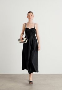Robe maxi noire sans manches avec un bodice froncé et des bretelles ajustables. Le modèle tient un sac à main rayé et porte des sandales plates noires.