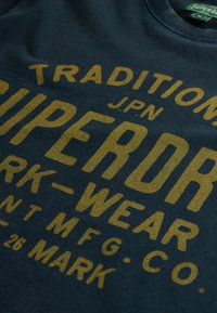 Mørkeblå t-shirt med store gyldne bogstaver, der står "SUPERDRY J.P.N TRADITIONAL WORK-WEAR." Blødt bomuldsstof med rund halsudskæring.