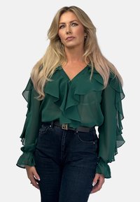 Green ruffled blouse with sheer fabric, v-neckline, button closure, and long sleeves. Paired with dark denim jeans and a belt.