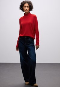 Roter Longsleeve-Pullover mit Rollkragen, kombiniert mit dunkelblauen weit geschnittenen Jeans. Das Outfit hat eine glatte Textur und eine figurbetonte Silhouette.