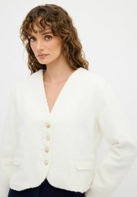 Chaqueta blanca texturizada con escote en forma de V, que presenta tres botones de oro y perlas, y dos bolsillos frontales. Tela suave y diseño corto.