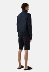 Veste bombardier marine avec un col montant, des poignets et une taille côtelés ; associée à un short noir et des sandales à glissière noires. Texture de tissu lisse.