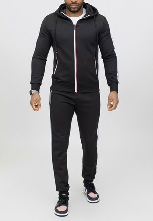 Haut à zip noir avec capuche à cordon, doté d'accents en rayures rouges et bleues. Associé à un jogging noir assorti, tous deux en tissu.