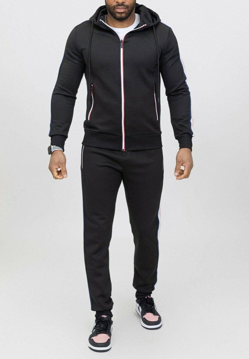 Haut à zip noir avec capuche à cordon, doté d'accents en rayures rouges et bleues. Associé à un jogging noir assorti, tous deux en tissu.