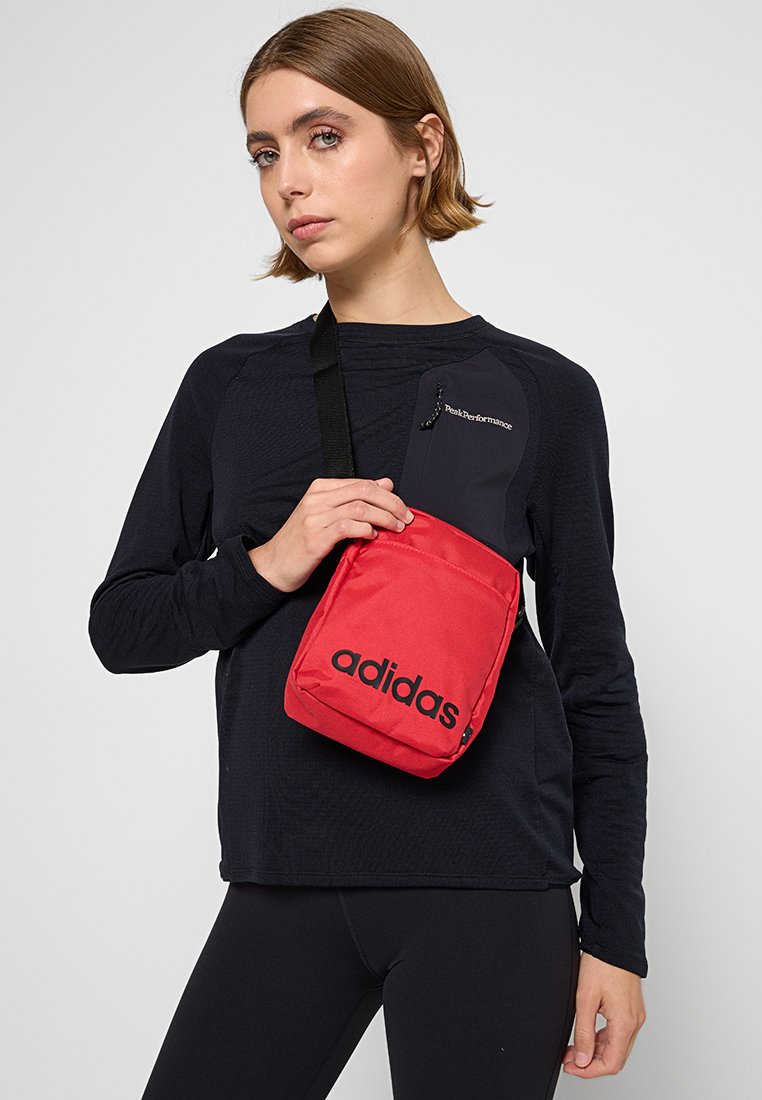 Bolsa bandolera roja de adidas con correa negra ajustable, de forma rectangular, con logotipo en la parte frontal y tejido texturizado. Llevada por un modelo en ropa deportiva.