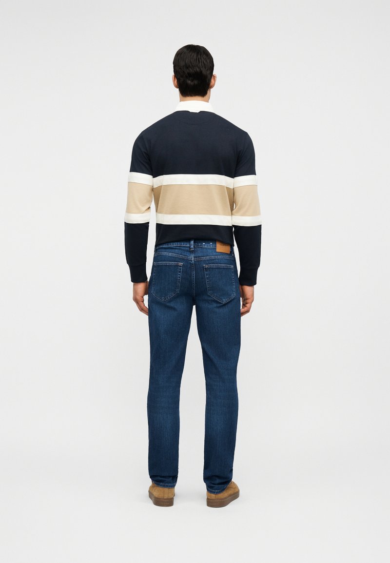Homme debout de dos, portant un pull à rayures bleu marine, beige et blanc, un jean bleu et des chaussures marron, sur un fond uni.