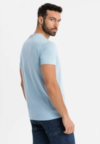 Světle modré tričko s krátkým rukávem vyrobené z bavlny, s volným střihem a hladkým povrchem. Model má na sobě tmavé džíny.