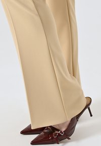 Beige Hosen mit weicher Textur und geradem Schnitt, kombiniert mit glänzenden burgunderfarbenen Absatzschuhen mit spitzem Zehenbereich, die über ein doppeltes Schnallen-Detail verfügen.