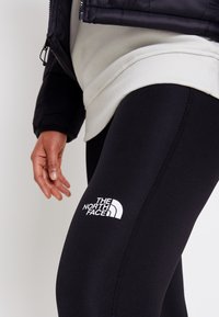 Czarne legginsy wykonane z elastycznego materiału, z białym logo "The North Face" na udzie i gładką fakturą.