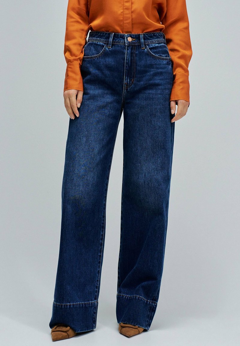 Femme portant un jean large bleu foncé, un blouse à manches longues orange et des chaussures à talons pointus marron sur un fond uni.