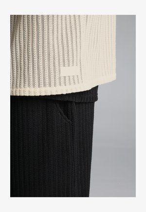 Top leggero in maglia color crema con un motivo testurizzato, abbinato a pantaloni neri a coste con tasca. Etichetta visibile sull'orlo superiore.