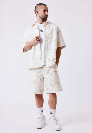 Homme portant une chemise blanche à manches courtes à motifs et boutons, un short assorti, un t-shirt blanc avec un motif, des baskets blanches et des chaussettes blanches, debout et regardant vers la gauche.