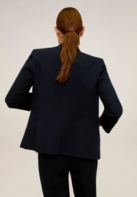 Femme aux cheveux roux attachés en queue de cheval, portant un blazer noir et un pantalon noir, debout les mains sur les hanches, face à un fond beige uni.