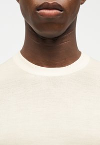 Κοντινό πλάνο του κάτω μέρους του προσώπου και του λαιμού ενός σκούρου δέρματος ατόμου που φοράει ένα απλό, εκρού μπλουζάκι με στρογγυλή λαιμόκοψη, με φόντο λευκό.