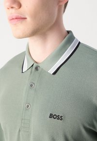 Fiatal férfi világoszöld pólót visel, fehér és fekete csíkos gallérral, a mellkasán pedig a "BOSS" logó hímzett.