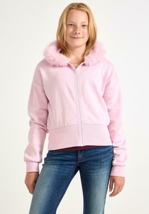 Ung person iført en lyserød lynlåstrøje med en fluffy hætte, blå jeans, og står mod en hvid baggrund.
