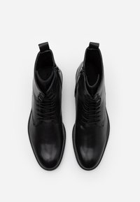 Botas de tobillo de cuero negro con una textura suave, que presentan cordones y un cierre con cremallera lateral. Puntera redondeada y detalles de costura.