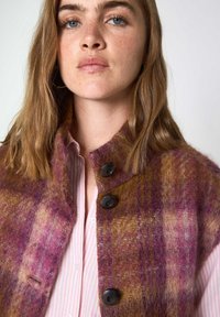 Gilet en mélange de laine dans des teintes de violet et de brun, avec un col haut, quatre boutons noirs et un motif texturé à carreaux.