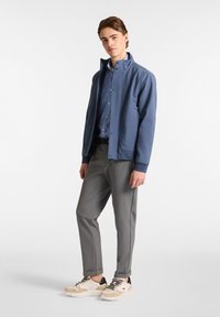 Chaqueta azul de concha blanda con cuello alto, pantalones grises y zapatillas beige; presenta un cierre de botones en el frente y dos bolsillos laterales.
