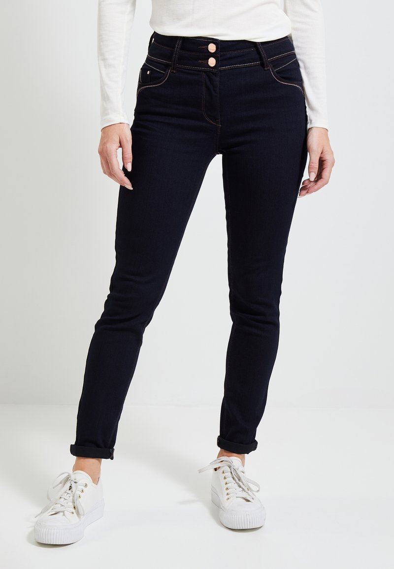 Jean en denim bleu foncé à taille haute, fermeture à double bouton et manches retroussées ; associés à des baskets blanches avec œillets dorés.
