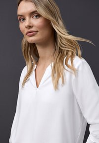 Blusa bianca a maniche lunghe con scollo a V. Tessuto morbido e liscio. Il modello ha capelli castano chiaro con morbide onde e uno sfondo neutro.