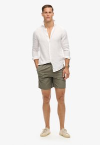 Camicia bianca di lino con maniche arrotolate, abbinata a pantaloni corti verde oliva. Sneakers beige chiaro completano il look, mostrando uno stile casual.