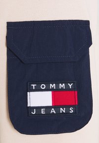 Marinblå tygficka med ett rektangulärt lock som har en broderad "TOMMY JEANS"-etikett och en tri-färgad logo i rött, vitt och blått.