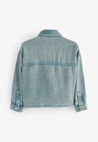 Jasnoniebieska kurtka dżinsowa z długimi rękawami i srebrnymi ćwiekami ułożonymi w pionowe linie na górnej części pleców i ramionach.