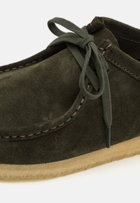 Vihreä mokkanahkasaapas, jossa on pyöristetty kärki, tikattu yksityiskohta ja vastaavasta vihreästä kankaasta valmistettu nauhakiinnitys. Pohja on vaaleanruskea.