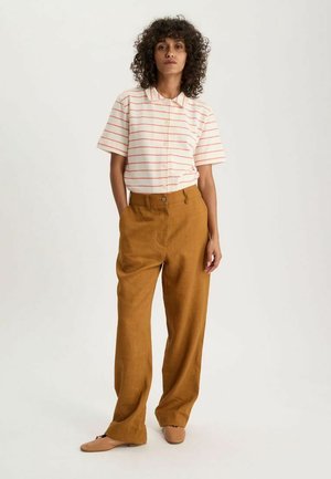 Femme aux cheveux bouclés portant un t-shirt blanc à manches courtes avec des rayures rouges, un pantalon large couleur moutarde et des ballerines beiges, debout les mains dans les poches.