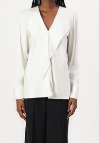 Blusa bianca a maniche lunghe con scollatura a V profonda e dettaglio frontale a strati. Tessuto morbido con una consistenza delicata, abbinata a pantaloni neri.