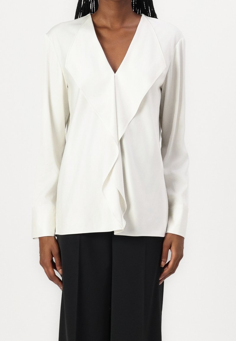 Blusa bianca a maniche lunghe con scollatura a V profonda e dettaglio frontale a strati. Tessuto morbido con una consistenza delicata, abbinata a pantaloni neri.